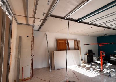 Installation du placo au plafond, avec gaine électrique et ossature de rails pour le faux plafond visibles. Lève-plaque en action sur le chantier.