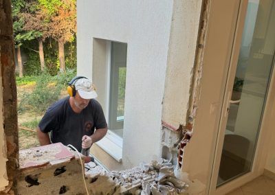 Démolition d'un mur pour préparer l'installation d'une baie vitrée, avec des outils de chantier visibles et des débris de construction.