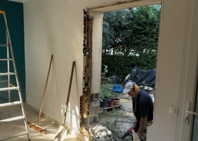 Démolition d'un mur pour préparer l'installation d'une baie vitrée, avec des outils de chantier visibles et des débris de construction.
